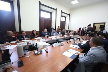 Personas sentadas alrededor de una mesa de madera con micrófonos, carpetas, botellas de agua y rótulos de identificación durante una reunión