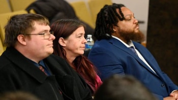 Tres personas sentadas en una sala de tribunal. Un hombre con gafas, una mujer con cabello rojizo y un hombre con barba y dreadlocks observan la escena