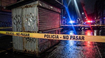 Un puesto metálico cerrado tras una cinta amarilla de policía, con vehículos policiales e iluminación de sirenas en una calle nocturna.