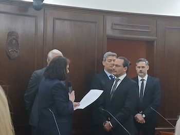Una mujer con micrófono lee un papel frente a un hombre de traje, con otras tres personas formadas al fondo en una sala de madera