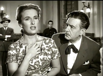 Fotograma en blanco y negro de una mujer con vestido estampado y la mano en el pecho junto a un hombre con traje y corbata de lazo