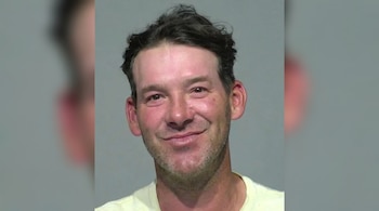 Un hombre con cabello oscuro y barba incipiente, vistiendo una camiseta de color claro, posa frente a un fondo gris.