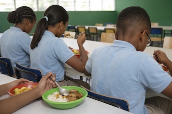 Vista trasera de varios niños con uniformes celestes comiendo de platos en un comedor escolar con mesas y sillas de fondo.