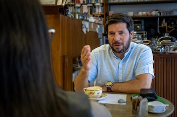 Hombre barbudo con camisa clara y reloj amarillo gesticula en una mesa con taza de café, papel, bolígrafo y servilletas frente a una mujer de espaldas