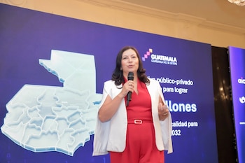 Una mujer con traje rojo y saco blanco habla por micrófono frente a un mapa de Guatemala y una pantalla azul