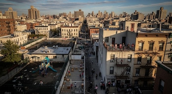 Vista aérea de un barrio denso de la ciudad con edificios, un patio de juegos de colores, personas en la calle y un paisaje urbano de fondo.