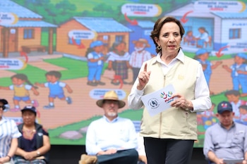 Una mujer con chaleco beige sostiene una tarjeta del programa Mano a Mano mientras habla ante un mural ilustrado y personas sentadas