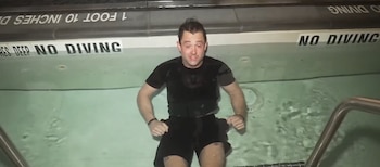 Un hombre caucásico de pelo oscuro y camisa negra está en una piscina con agua clara, con señales de "NO DIVING" en el borde y barandillas metálicas a la derecha