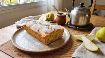 Un budín espolvoreado con azúcar en un plato, peras frescas, un mate con bombilla y una pava metálica sobre una mesa de madera.