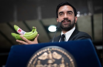 Hombre con barba y traje sostiene plátanos verdes con etiqueta de descuento del 30% y código QR, hablando en un micrófono detrás de un podio azul