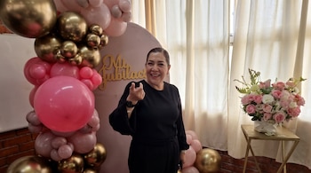 Una mujer vestida de negro gesticula con la mano frente a una columna de globos, un letrero de madera y un florero sobre una mesa.