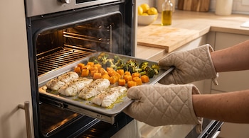 Horno abierto con vapor, bandeja de metal con cuatro filetes de merluza, cubos de calabaza y ramilletes de brócoli asados, dos manos con guantes de horno.