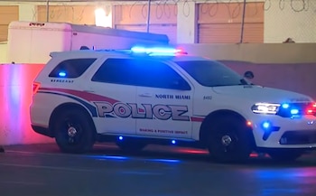 SUV policial blanco de North Miami con luces azules y rojas intermitentes estacionado en la oscuridad, con un remolque y una cerca al fondo