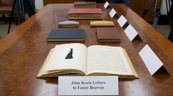 Libro antiguo abierto en una mesa de madera con caligrafía y silueta femenina. Otros libros cerrados y carteles blancos forman parte de la exposición.