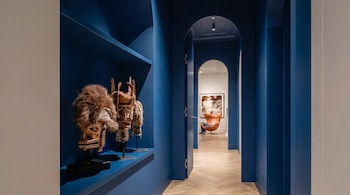 Pasillo interior con paredes azul cobalto, estante con tres máscaras tribales decorativas y dos arcos que conducen a una habitación con un sillón naranja.