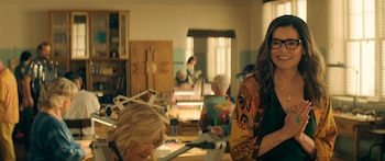 Geena Davis sonriendo con gafas y una chaqueta estampada en un taller. Detrás de ella, otras personas trabajan en mesas con herramientas y estanterías