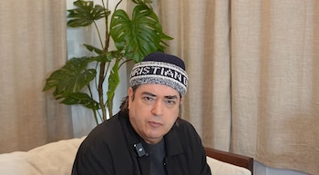 Jaime Bayly con gorro tejido impreso y chaqueta oscura, sentado frente a una planta de interior y cortinas de tono claro