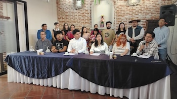 Grupo de personas en una sala con mesa, mantel azul, micrófono, vasos, papeles, pared de ladrillo y altavoz negro