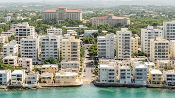 Plano general aéreo de una ciudad costera con edificios residenciales modernos y casas bajas frente al mar, con hoteles al fondo. Se aprecian letreros de alquiler.