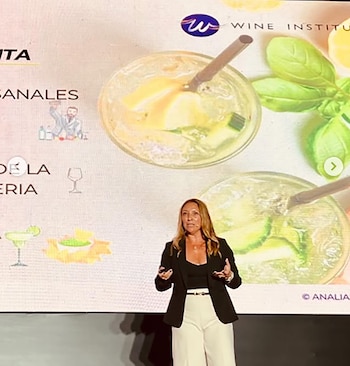 Mujer de cabello largo en blazer negro y pantalón blanco, de pie frente a una pantalla con imágenes de bebidas con hielo, fruta y hojas verdes