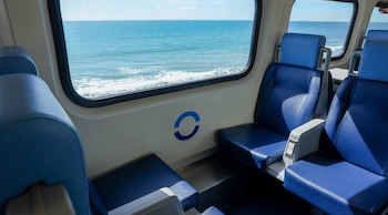 Interior de un vagón de tren con asientos azules y paredes blancas. Ventana con vista al océano, olas. Logo azul circular en pared y tomas de corriente