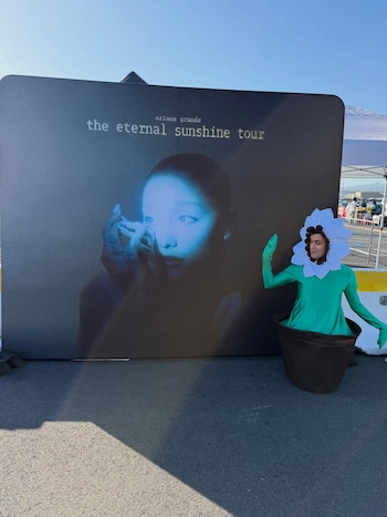 Giacomo Benavides, disfrazado de flor saliendo de una maceta, posa junto a un cartel de Ariana Grande para su gira 'The Eternal Sunshine Tour'