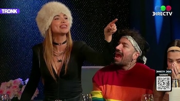 Una mujer con sombrero de piel, un hombre con bandana y suéter de rayas, y una tercera persona con manos en la boca, sentados a una mesa