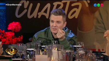 Hombre con chaqueta verde sentado a una mesa con vasos, tazas, velas encendidas. Fondo oscuro con texto "CUARTA" y logo ojo dorado. Hashtag Gran Hermano