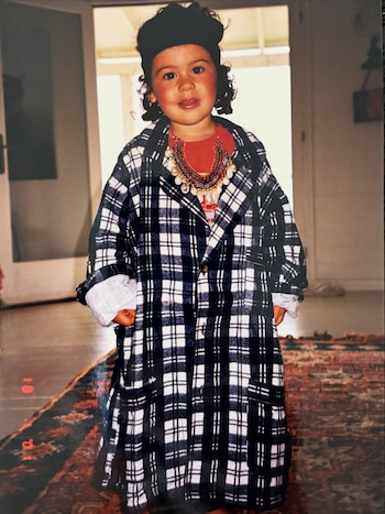 Una niña pequeña posa de pie sobre una alfombra con un abrigo holgado a cuadros blancos y negros, collares en el cuello y un gorro oscuro