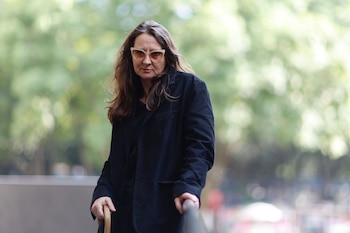 Lucrecia Martel, de mediana edad, con cabello castaño, gafas de sol y chaqueta negra, sostiene un bastón y un pasamanos, mirando al frente con un fondo verde desenfocado