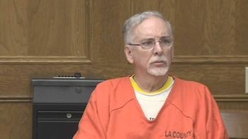 James M. Heaps, un hombre mayor con barba gris y gafas, viste un uniforme de cárcel naranja con "LA COUNTY" visible, sentado ante un panel de madera