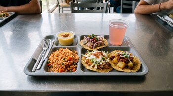 Charola gris con arroz, tres tostadas, postre, cubiertos y un vaso con bebida rosa sobre una mesa de acero inoxidable.