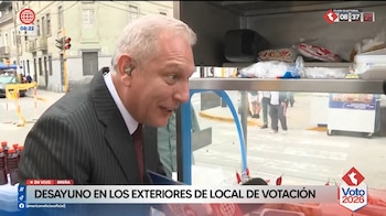 Gunter Rave, un hombre de cabello blanco con traje y corbata, se inclina hacia un puesto de comida callejera con botellas de líquido rojo, en un entorno urbano