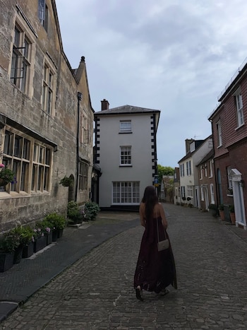Vista trasera de una mujer con un vestido largo color vino, caminando por una calle adoquinada flanqueada por edificios históricos de piedra y ladrillo, bajo un cielo nublado