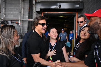 Tom Cruise sonriendo, con gafas de sol y polo negro, estrecha la mano de un hombre. Hay personas con acreditaciones y estructuras metálicas