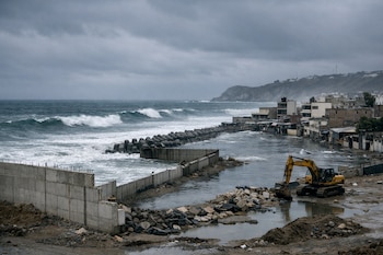 Cielo nublado sobre costa con mar agitado, olas grandes, dique de tetrapods, muros de concreto inconclusos, edificios costeros inundados y excavadora.