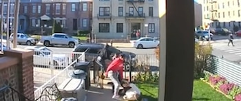 Imagen de CCTV mostrando a una persona de camiseta roja agrediendo a una anciana en el suelo en una acera de Brooklyn, junto a botes de basura y una valla blanca
