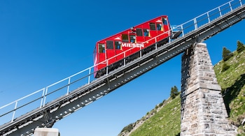 Funicular rojo Niesen subiendo por vía férrea metálica con barandales y pilares de piedra en ladera verde bajo cielo azul.