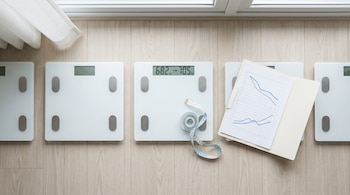 Varias básculas digitales blancas sobre un suelo de madera claro, una de ellas muestra 68.2 kg y 70.5 kg, junto a una cinta métrica y una carpeta con gráficos.
