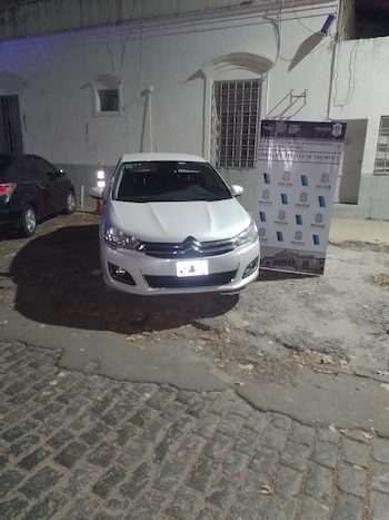 Un coche Citroën blanco estacionado de noche frente a la Comisaría Tercera de Quilmes, con un cartel policial visible al lado en una calle empedrada
