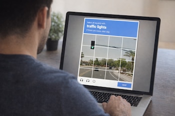 Una persona de espaldas sentada frente a una computadora portátil, en la pantalla se ve un CAPTCHA con imágenes de semáforos.