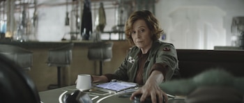 Mujer de mediana edad con chaqueta verde militar sentada en una mesa frente a un bar, con la mano extendida y un dispositivo en la mesa, Grogu se ve borroso al frente
