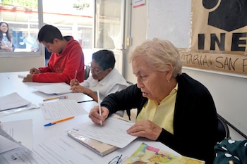 Tres personas, dos mujeres mayores y un hombre joven, sentadas en un escritorio blanco, escriben con lápices en hojas de papel durante una clase de alfabetización del INEA