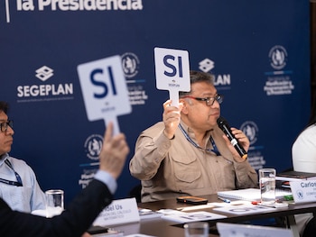 Hombres sentados en una mesa sostienen carteles blancos con 'SI' en azul. Uno habla por micrófono. Fondo azul con logo de SEGEPLAN