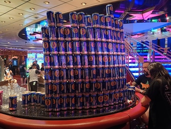 Una enorme torre de latas de cerveza Foster's de color azul y dorado apiladas sobre un mostrador de bar. Alrededor, personas, máquinas tragamonedas y luces de neón en un casino
