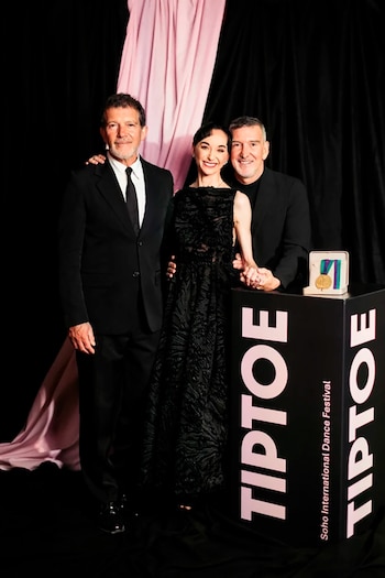 Tres personas sonríen frente a un podio negro con el logo "TIPTOE" y un premio dorado. Un hombre en traje y una mujer de vestido negro se toman de la mano, junto a otro hombre en traje oscuro