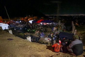 Vista aérea nocturna de un campamento con tiendas de campaña, personas reunidas, montones de ropa, botellas de agua. Al fondo se observa una ciudad iluminada