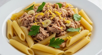 Plato blanco con fideos penne rigate cubiertos de atún desmenuzado, ralladura de limón, láminas de ajo y hojas de perejil fresco.