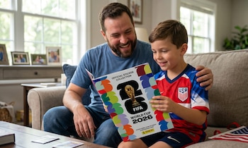 Un hombre barbudo sonríe junto a un niño en un sofá gris, ambos mirando un álbum de stickers del Mundial 2026. El niño lleva una camiseta de USA