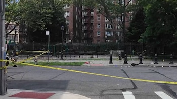 Vista de calle acordonada con cinta amarilla, con un oficial de policía examinando el suelo junto a una valla. Detrás hay un parque y edificios de apartamentos de ladrillo